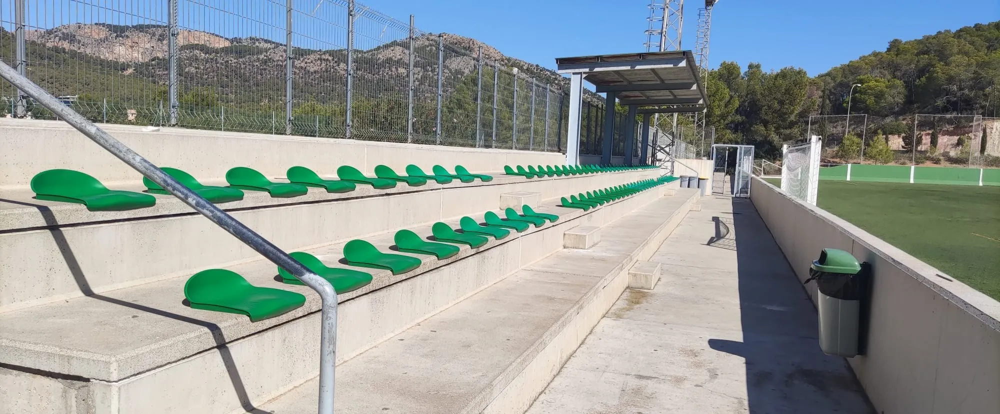 Grada de hormigón con asientos verdes y marquesina junto a campo de césped artificial