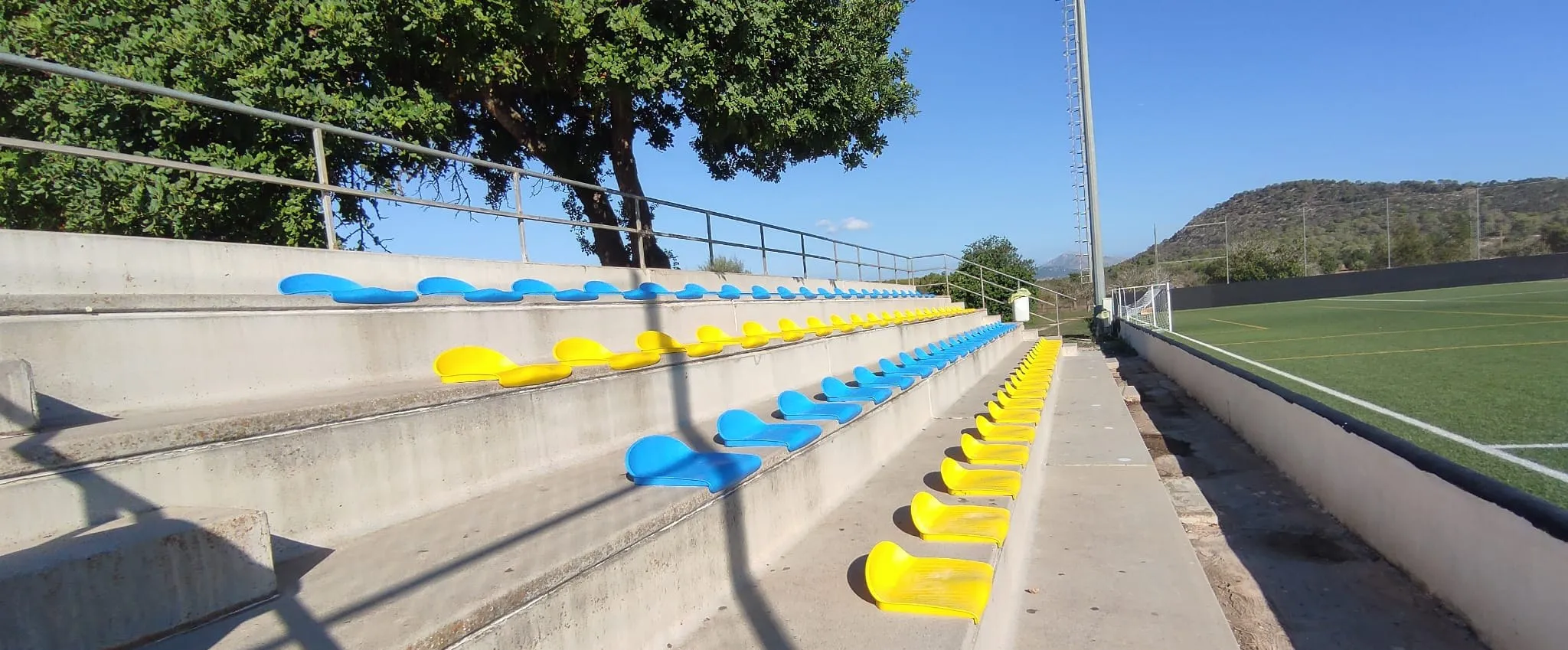 Grada de hormigón con asientos azules y amarillos recién instalados junto a un campo de fútbol