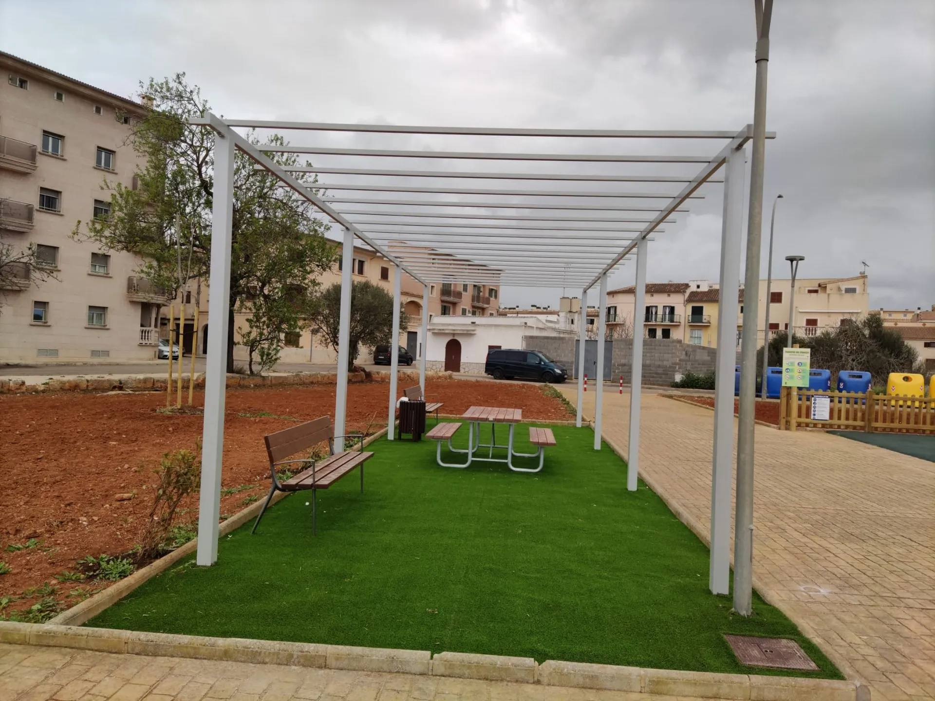 Pérgola metálica blanca sobre césped artificial con banco y mesa de pícnic