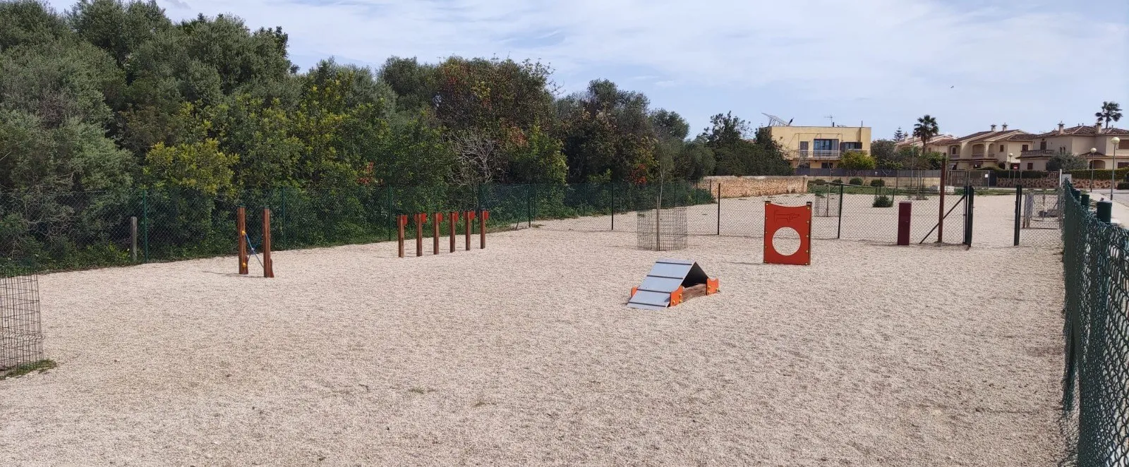 Recinto canino vallado con slalom, empalizada, panel de salto y árboles recién plantados sobre gravilla
