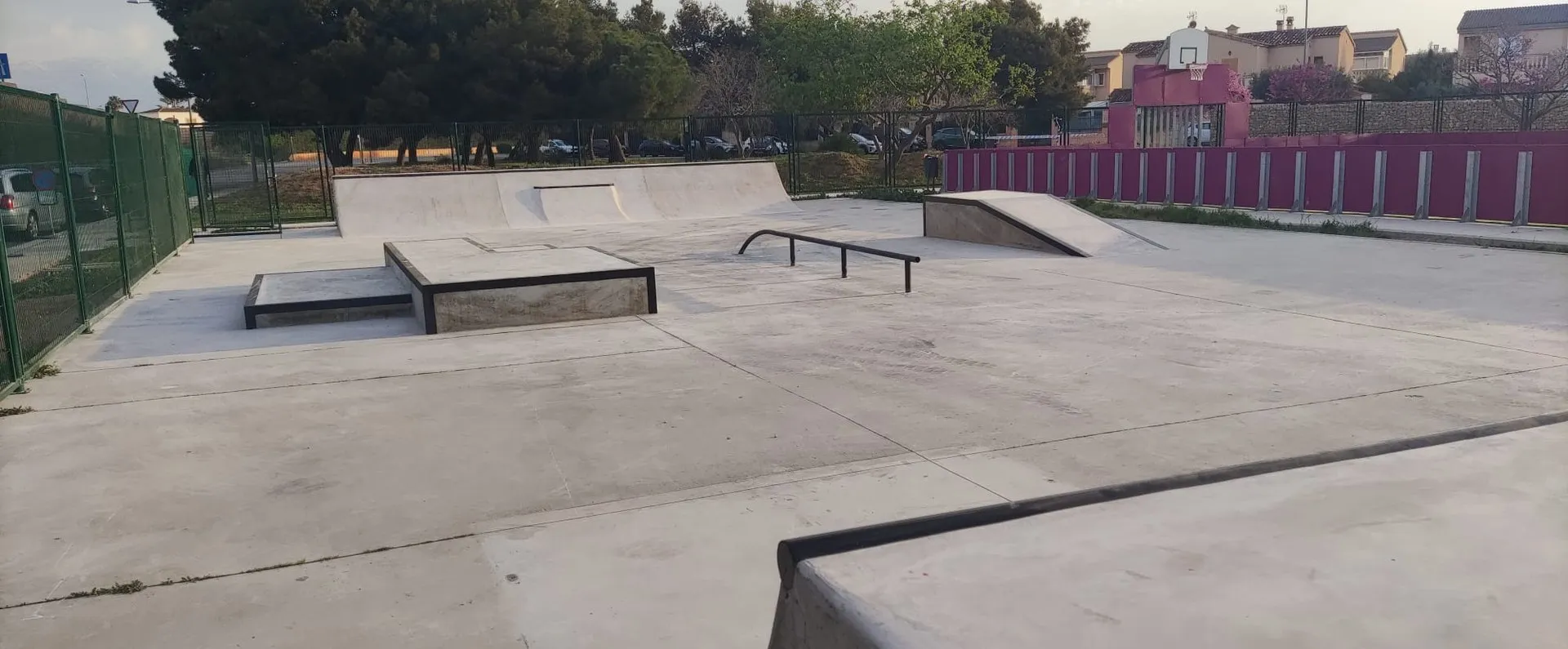 Skatepark de hormigón con módulos, rampa, cajón y barandilla junto a pista de baloncesto